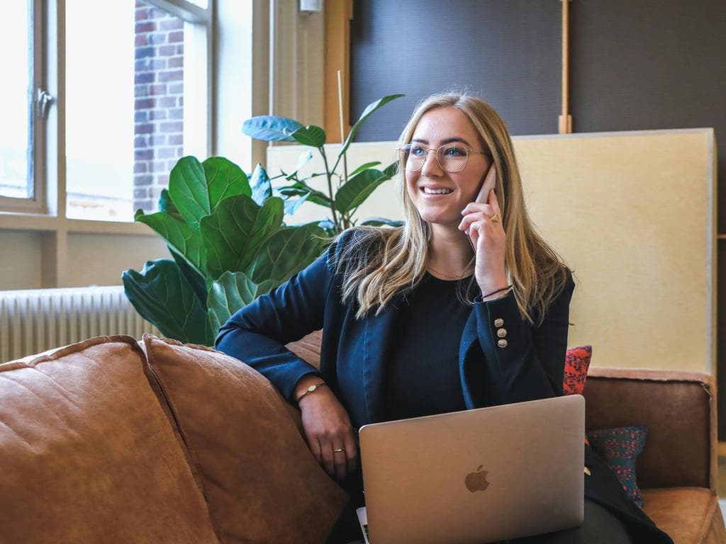 Woman sitting on couch making a call on her phone with her laptop on her lap, warm office setting.