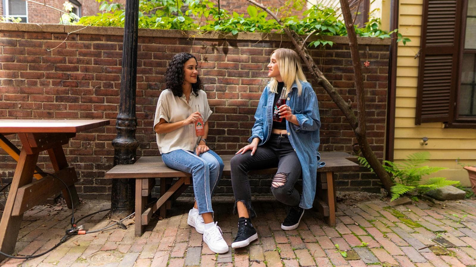 Two women sitting on a wooden brown bench and looking at each other smiling, each holding a soda bottle.